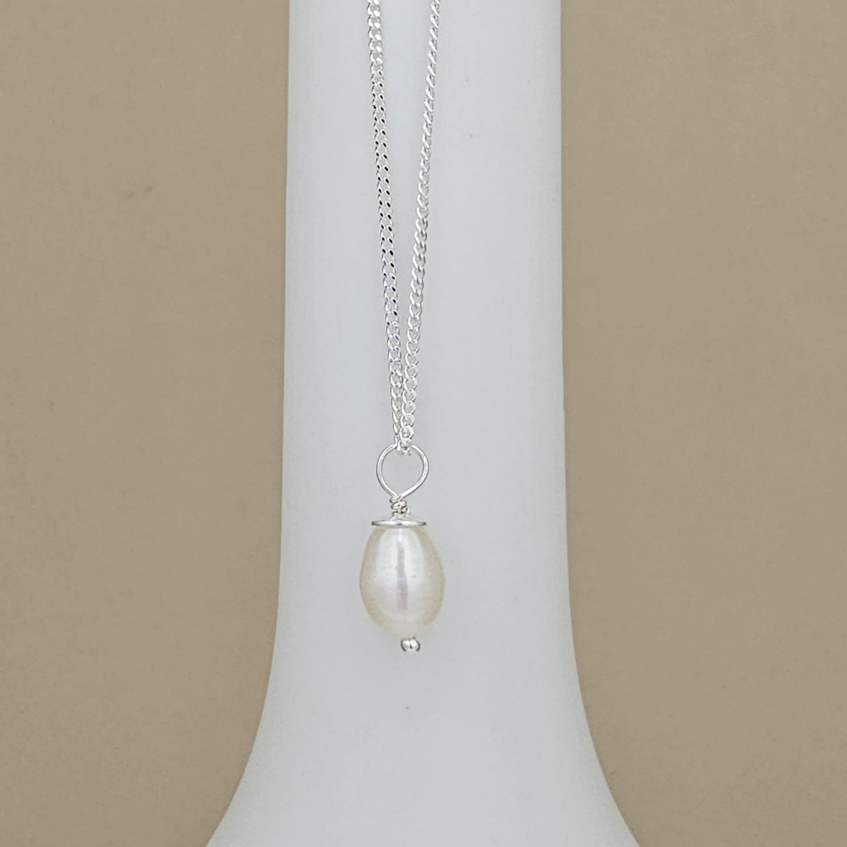 A silver chain necklace with a single teardrop-shaped white pearl pendant, displayed on a white jewelry bust against a beige background.