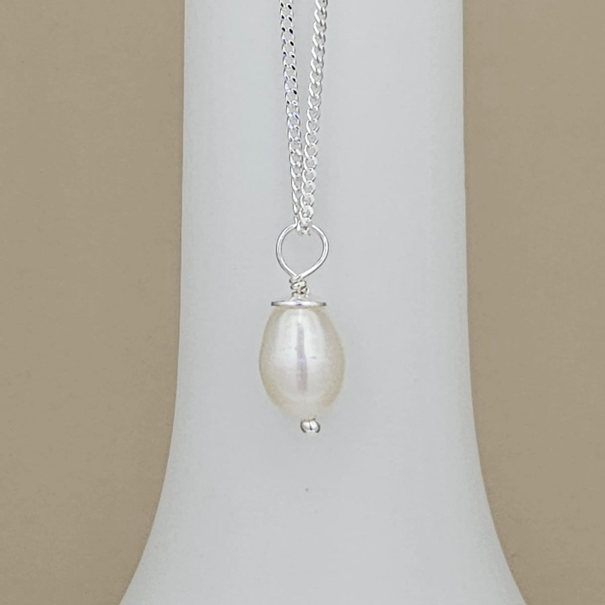 A silver chain necklace with a teardrop-shaped white freshwater pearl pendant, displayed on a white jewelry bust against a beige background.