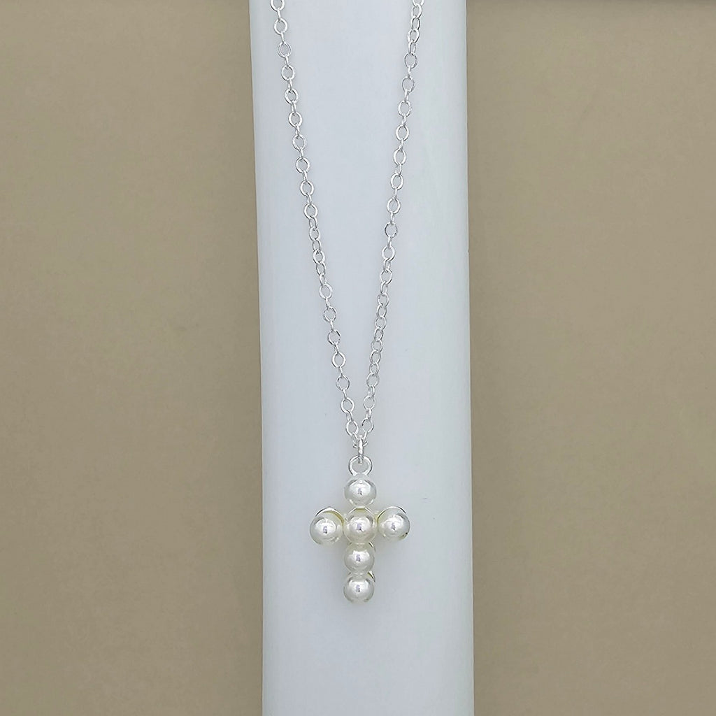 A silver chain necklace with a cross-shaped pendant made of white faux pearls, displayed on a white cylindrical stand against a beige background.