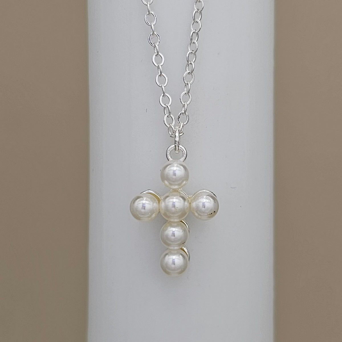 A silver chain necklace with a cross-shaped pendant made of five white pearl-like beads, displayed on a white cylindrical stand against a beige background.