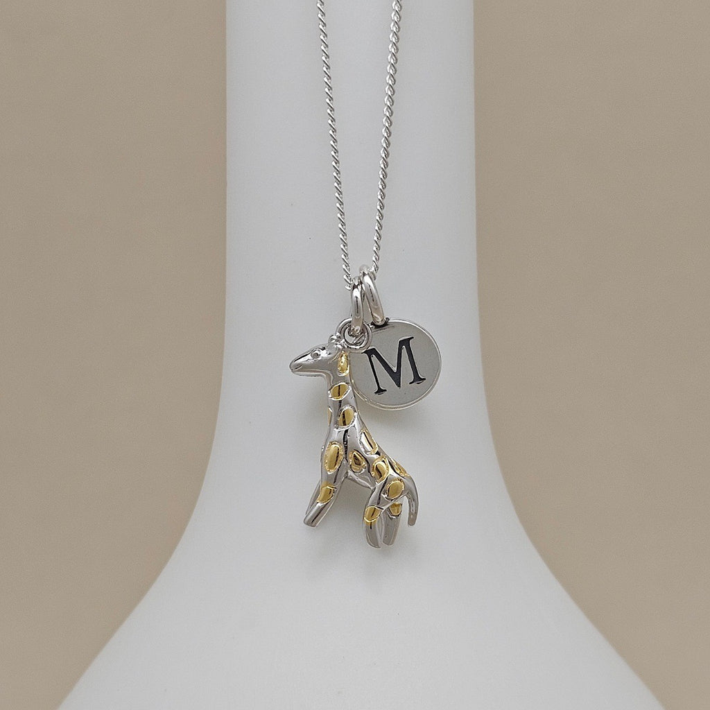 Necklace with a silver and gold giraffe pendant and silver disc featuring a letter 'M' on a white stand.