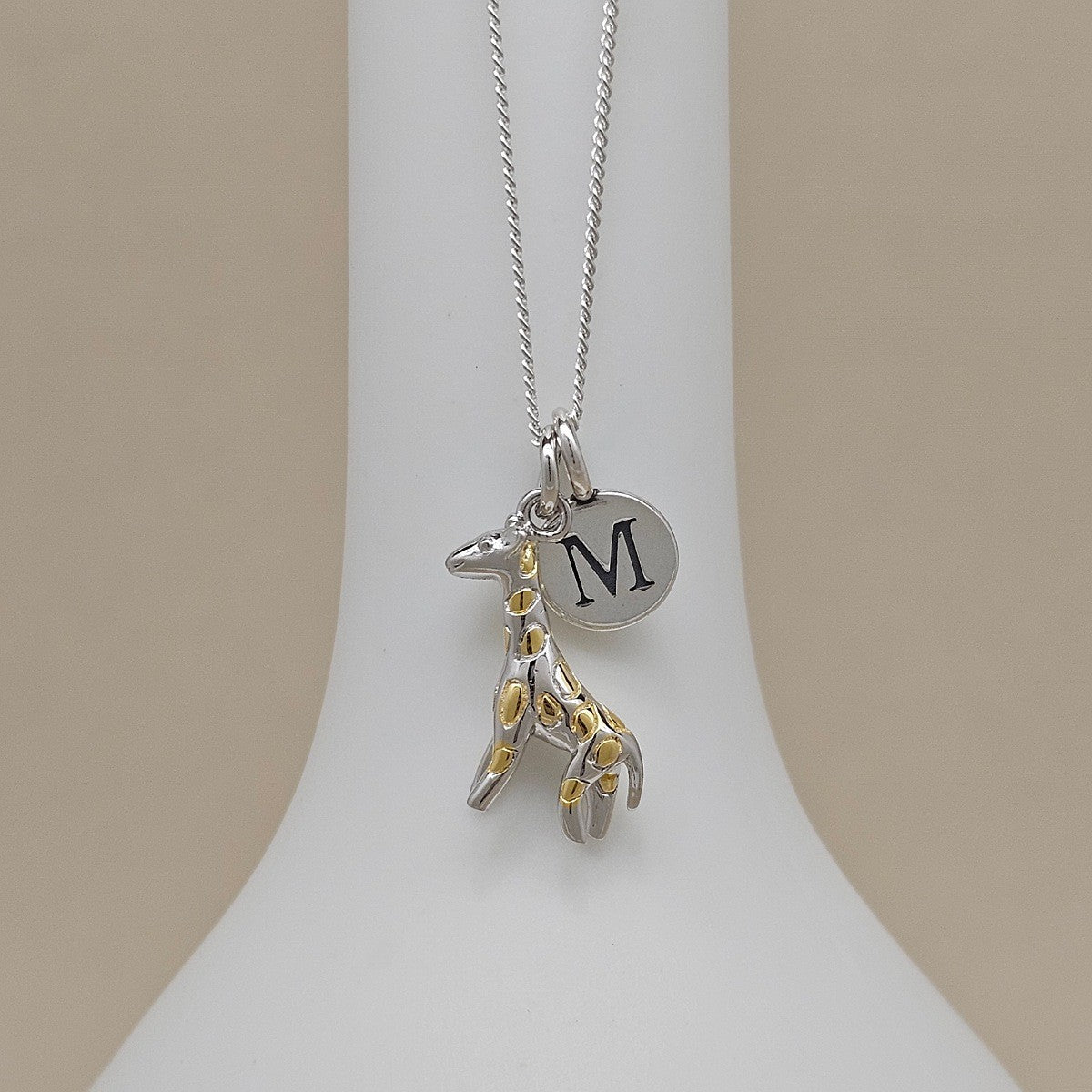 Necklace with a silver and gold giraffe pendant and silver disc featuring a letter 'M' on a white stand.