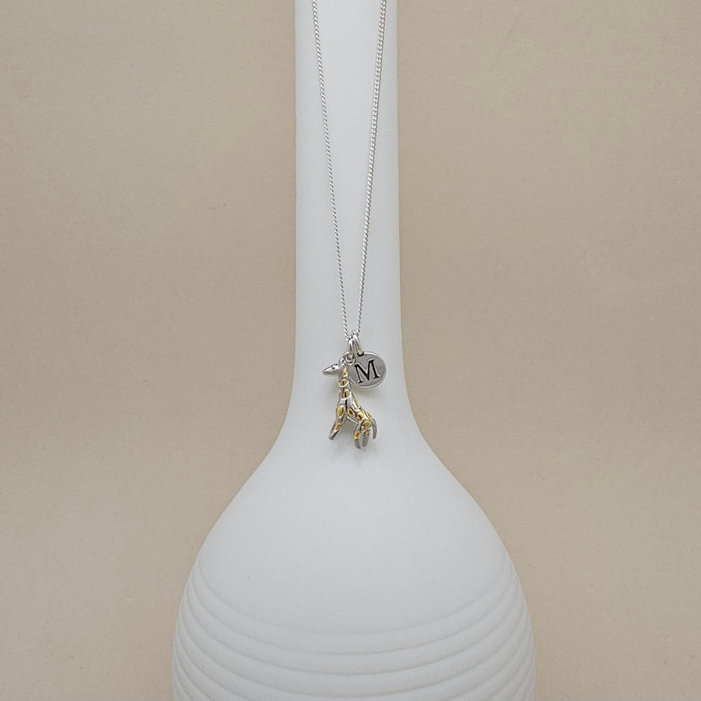 A silver necklace with a giraffe charm and a small round initial pendant engraved with the letter 'M', displayed on a white ribbed vase against a beige background.