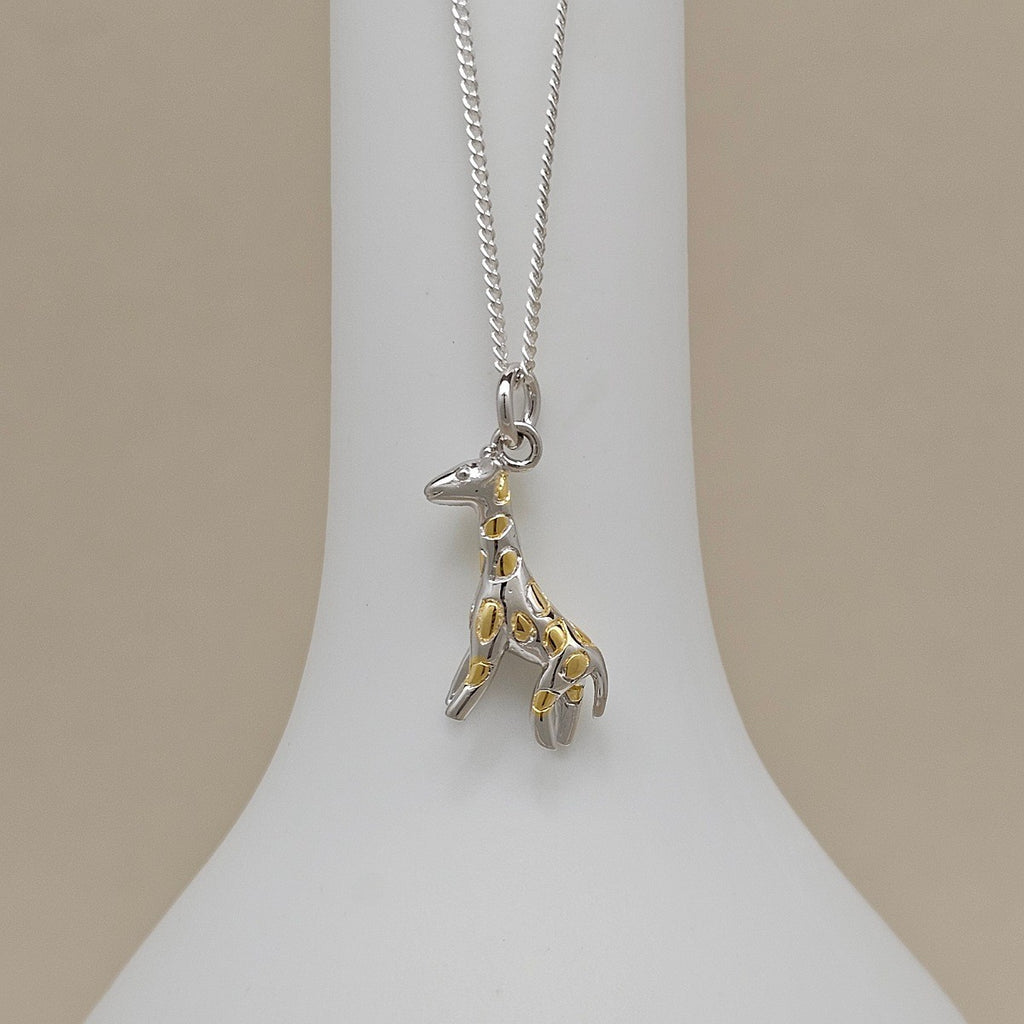 A silver giraffe pendant necklace with gold spot accents, displayed on a white neck stand against a beige background.