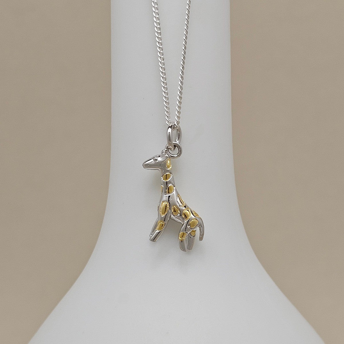 A silver giraffe pendant necklace with gold spot accents, displayed on a white neck stand against a beige background.