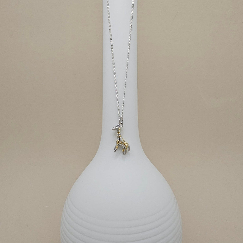 A delicate silver chain necklace with a small gold-toned giraffe pendant, displayed draped over a white ribbed ceramic vase against a warm beige background.