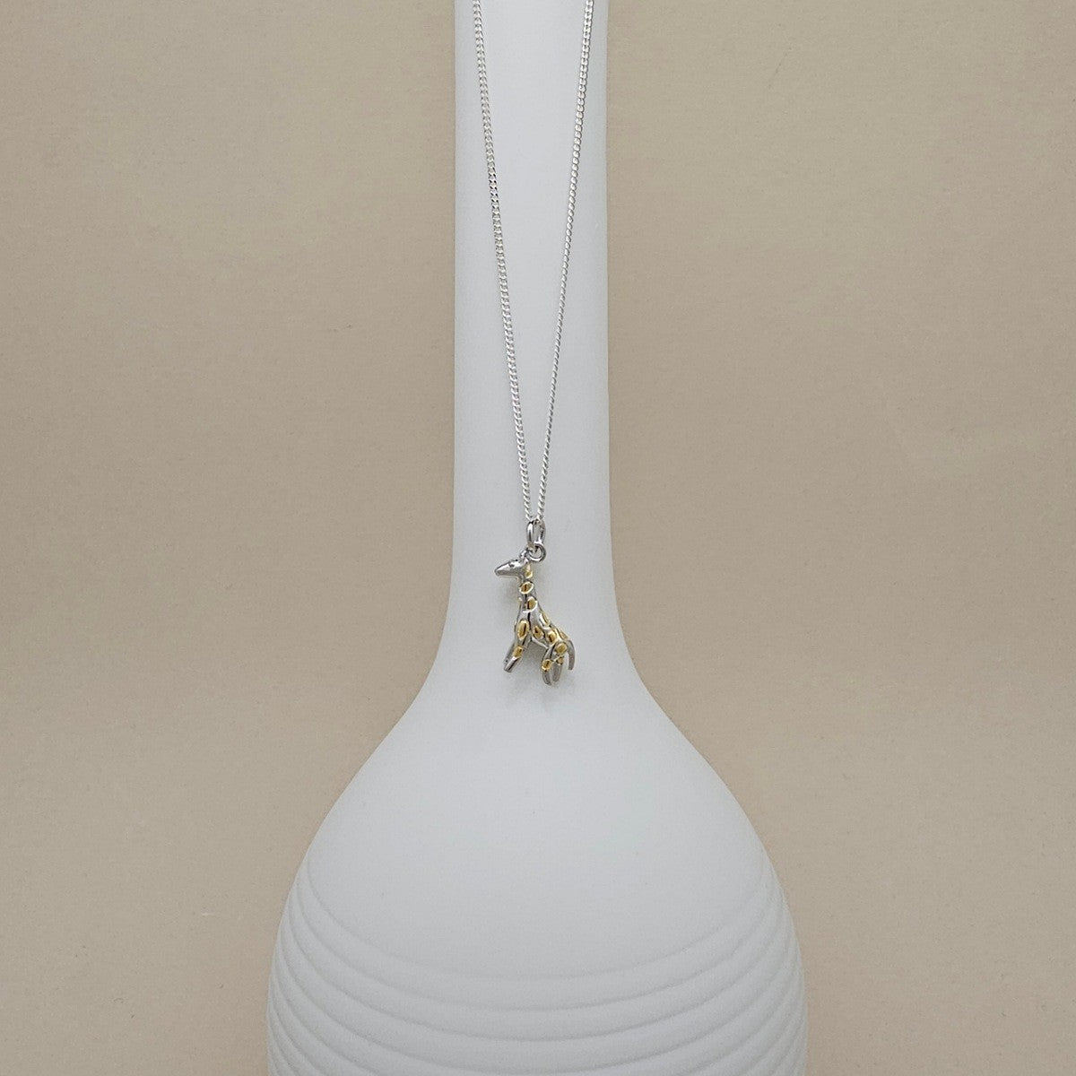 A delicate silver chain necklace with a small gold-toned giraffe pendant, displayed draped over a white ribbed ceramic vase against a warm beige background.