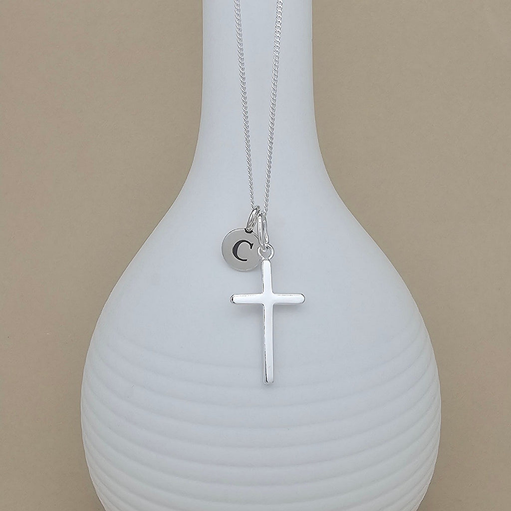 A silver necklace with a cross pendant and a small initial 'C' charm, displayed on a ribbed white vase against a beige background.
