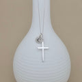 A silver necklace with a cross pendant and a small initial 'C' charm, displayed on a ribbed white vase against a beige background.