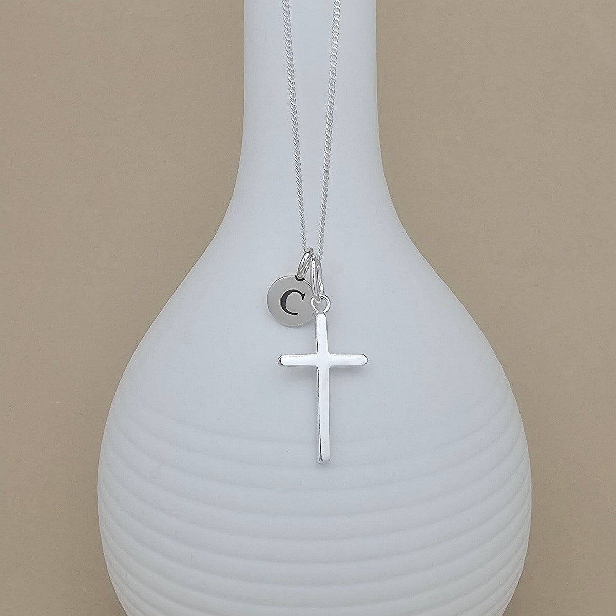 A silver necklace with a cross pendant and a small initial 'C' charm, displayed on a ribbed white vase against a beige background.