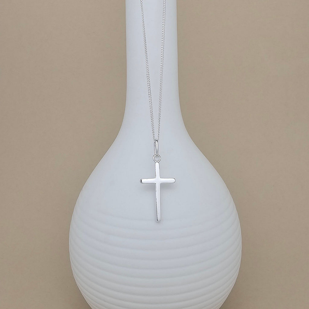 A silver cross pendant necklace hanging from a fine silver chain, displayed against a white ribbed vase on a warm beige background.