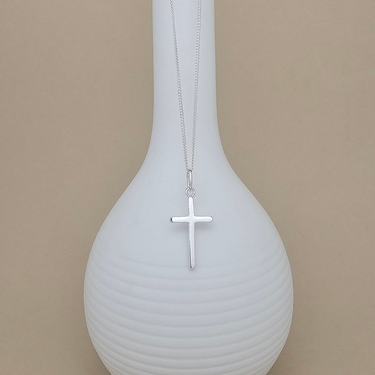 A silver cross pendant necklace hanging from a fine silver chain, displayed against a white ribbed vase on a warm beige background.