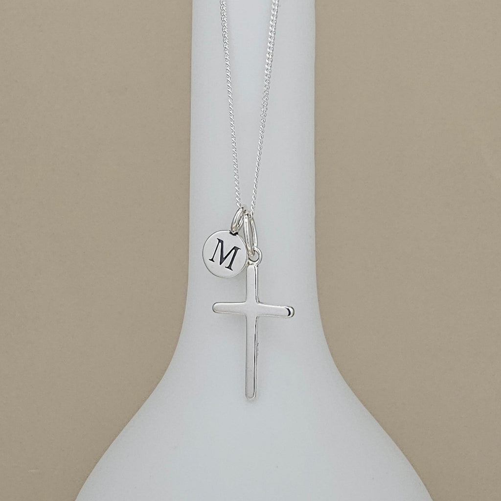 A silver cross necklace with a small circular initial 'M' charm, displayed on a white vase against a beige background.