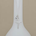 A silver cross necklace with a small circular initial 'M' charm, displayed on a white vase against a beige background.