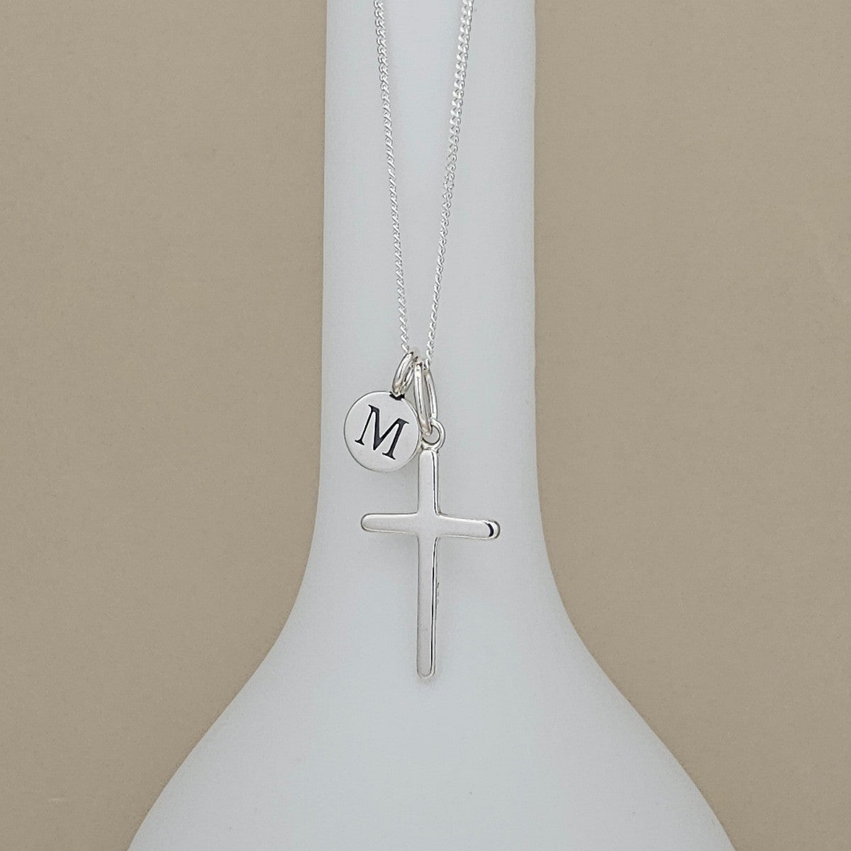 A silver cross necklace with a small circular initial 'M' charm, displayed on a white vase against a beige background.