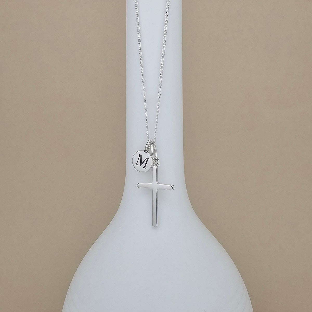 A silver necklace with a cross pendant and a small round initial charm engraved with the letter 'M', displayed on a white vase against a beige background.