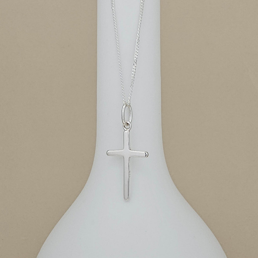 A silver cross pendant necklace hanging from a fine silver chain, displayed against a white vase-shaped stand on a beige background.