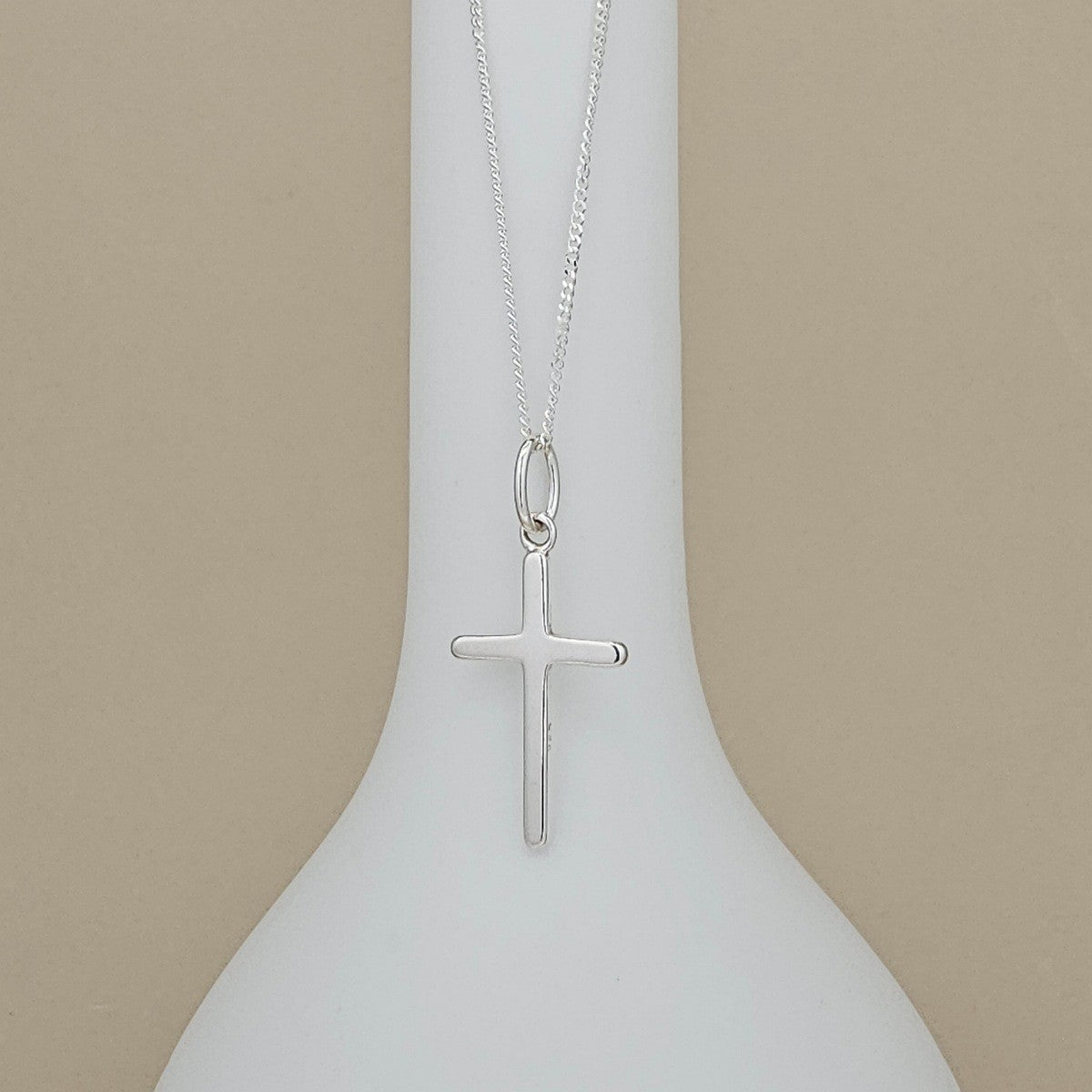 A silver cross pendant necklace hanging from a fine silver chain, displayed against a white vase-shaped stand on a beige background.