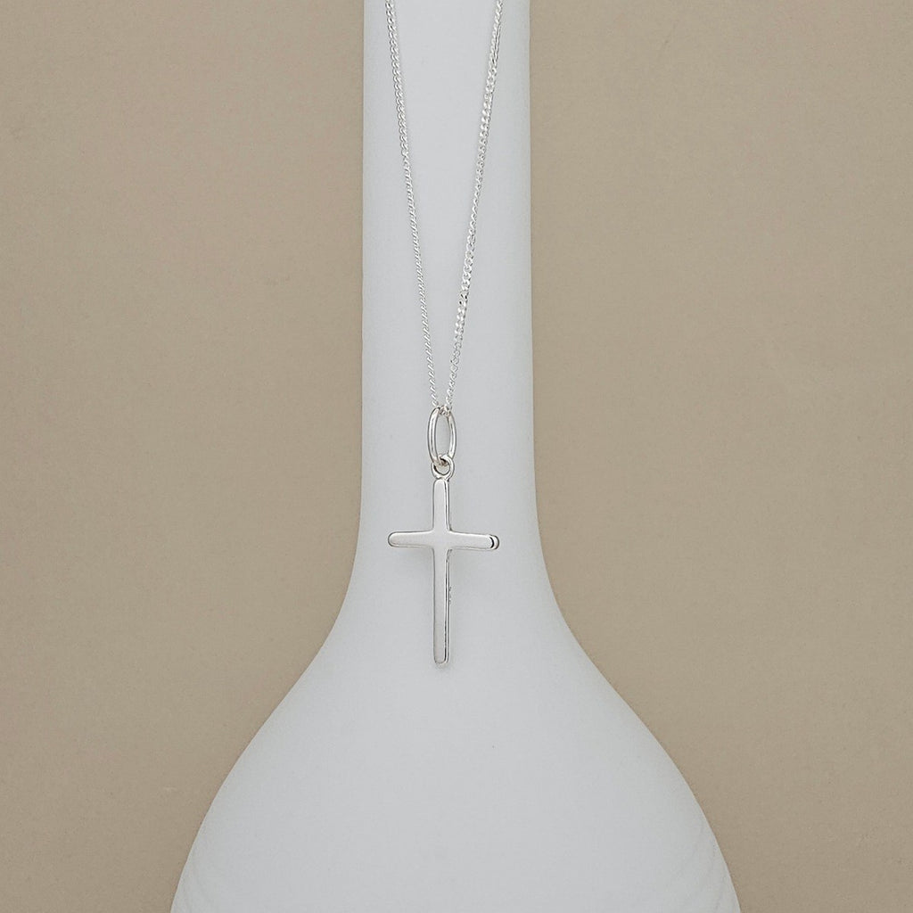 A silver cross pendant necklace hanging from a fine silver chain, displayed on a white ceramic vase against a warm beige background.