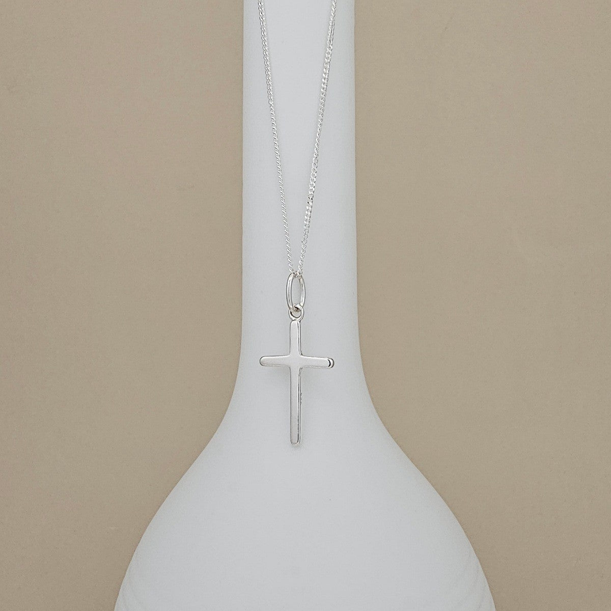A silver cross pendant necklace hanging from a fine silver chain, displayed on a white ceramic vase against a warm beige background.