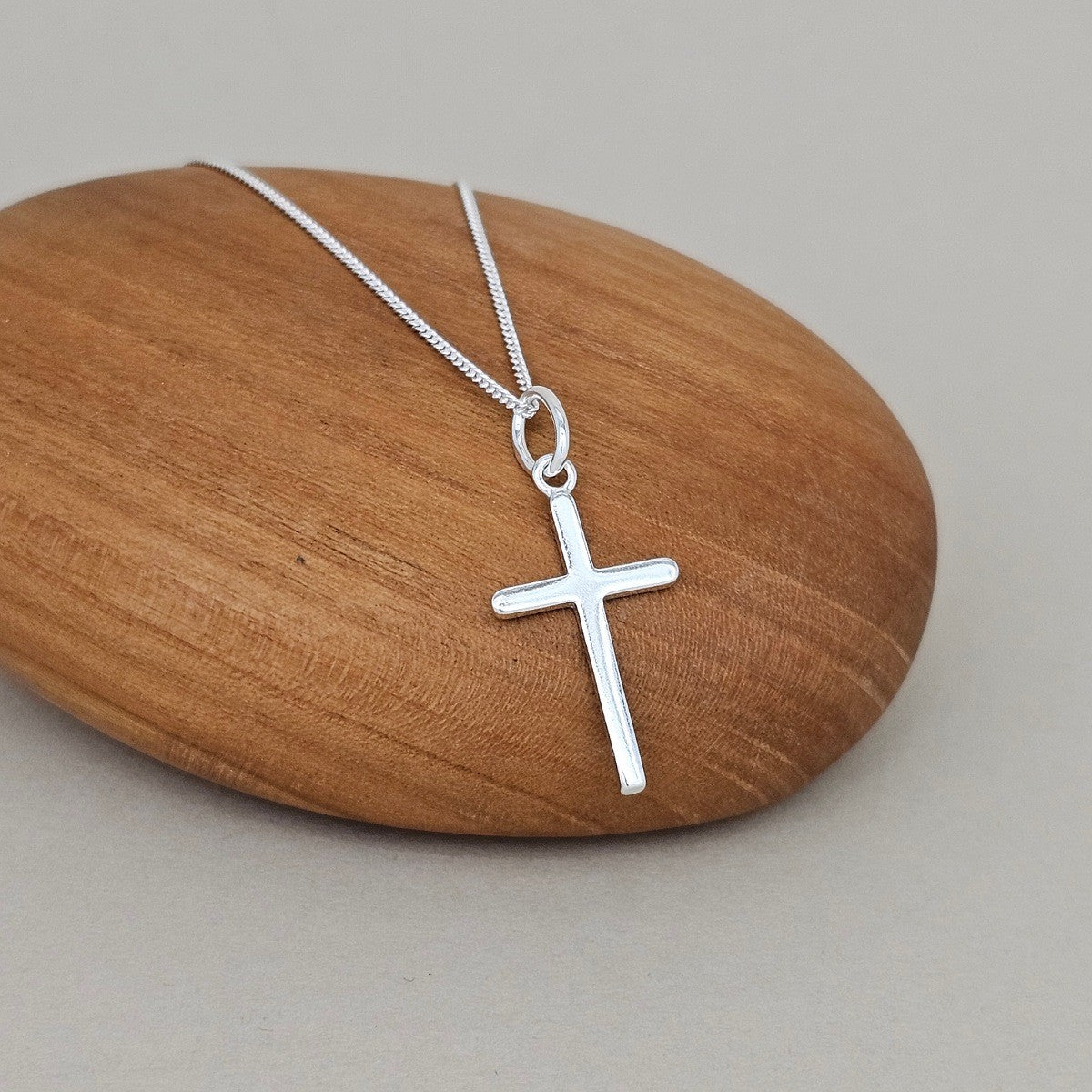 A silver cross pendant necklace on a delicate silver chain, displayed on a smooth, polished wooden stone against a neutral gray background.