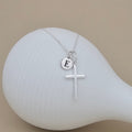 Silver necklace with a cross pendant and round initial charm on a white stand.