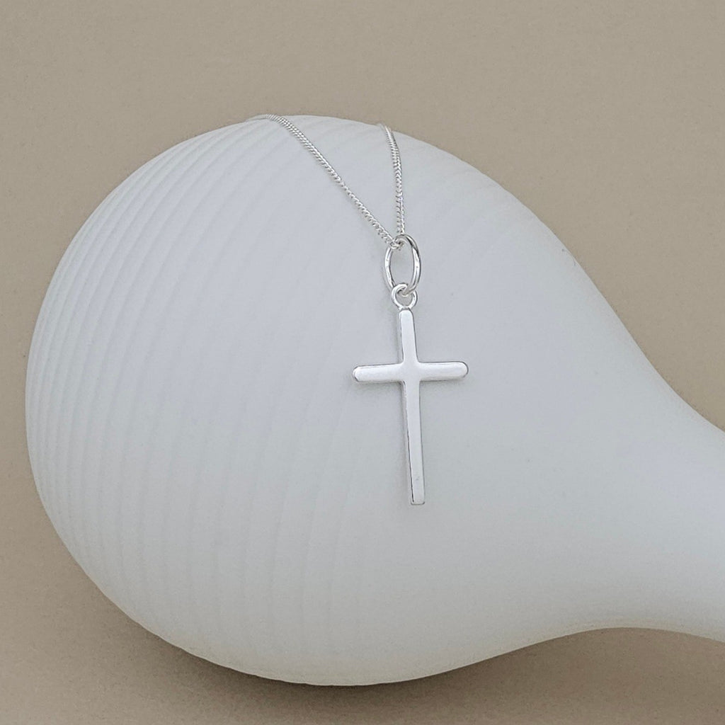 A silver cross pendant necklace on a delicate silver chain, displayed on a white ceramic mannequin against a warm beige background.