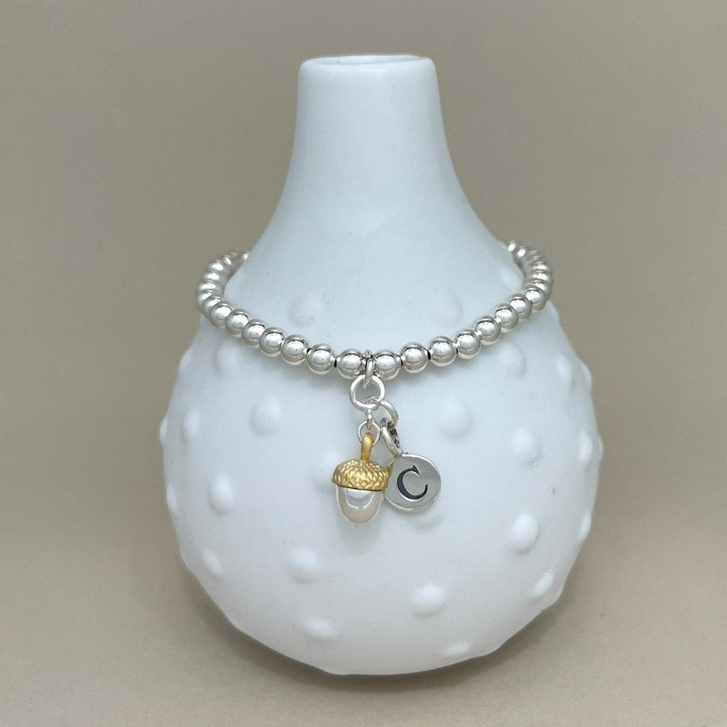 A silver beaded bracelet with a dangling acorn charm and a small circular letter C charm, displayed on a white textured ceramic vase against a neutral beige background.
