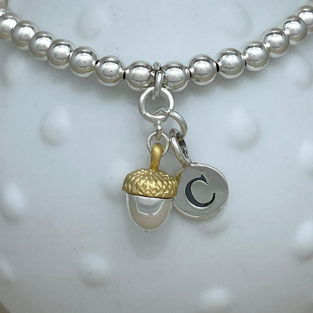A silver beaded bracelet with a dangling acorn charm and a small circular letter C charm, displayed on a white textured ceramic vase against a neutral beige background.