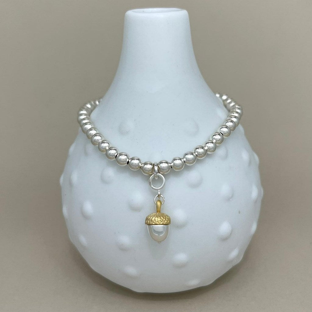 A silver beaded bracelet with a gold acorn pendant, displayed on a white textured ceramic vase against a neutral beige background.