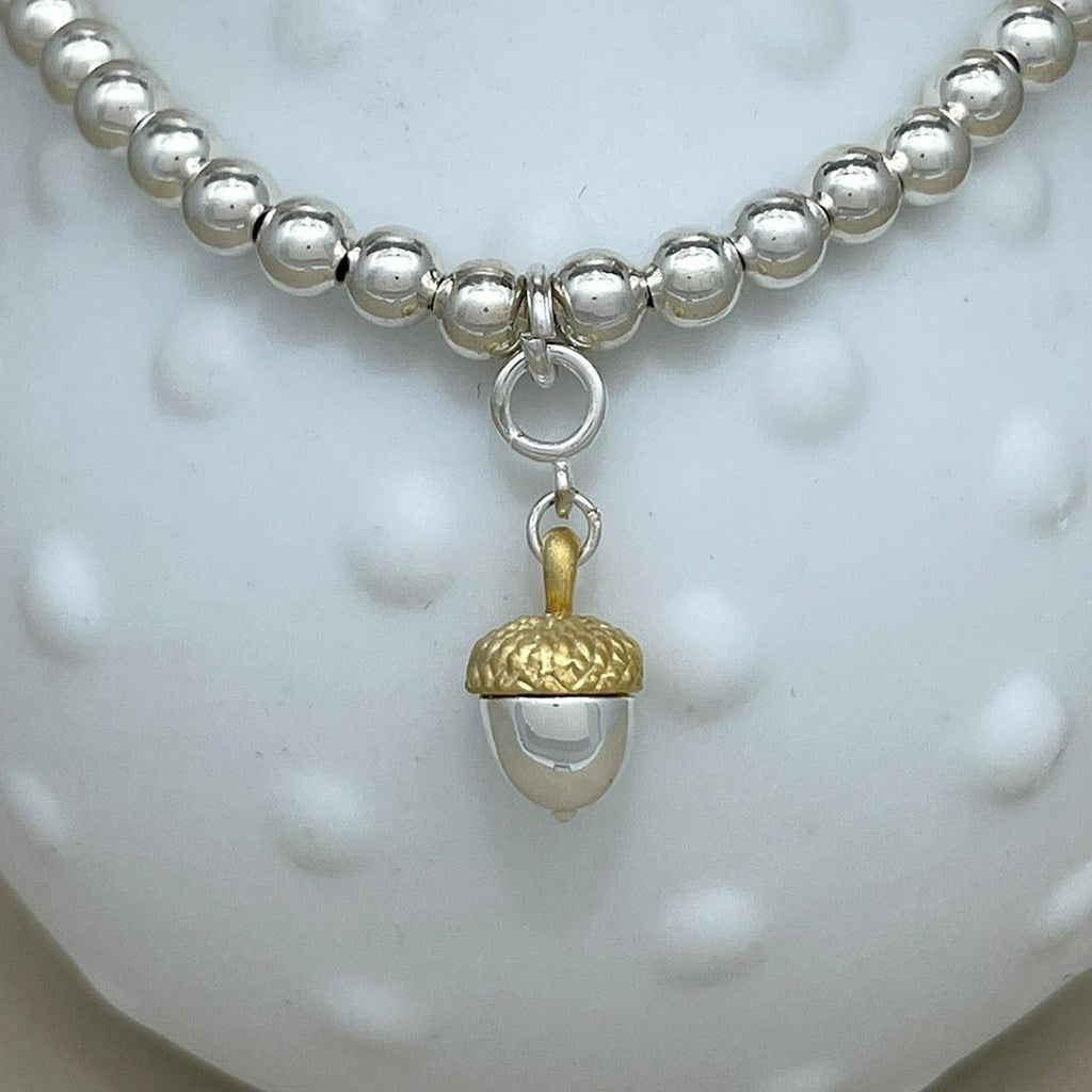 A silver beaded bracelet with a dangling acorn charm featuring a textured gold cap and a silver lower section, displayed on a white textured surface.