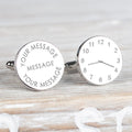 A pair of round silver cufflinks with one face showing a clock face design and the other showing customisable engraved text, set on a light wood background.