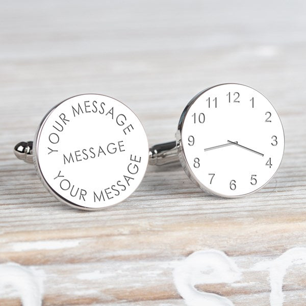 A pair of round silver cufflinks with one face showing a clock face design and the other showing customisable engraved text, set on a light wood background.