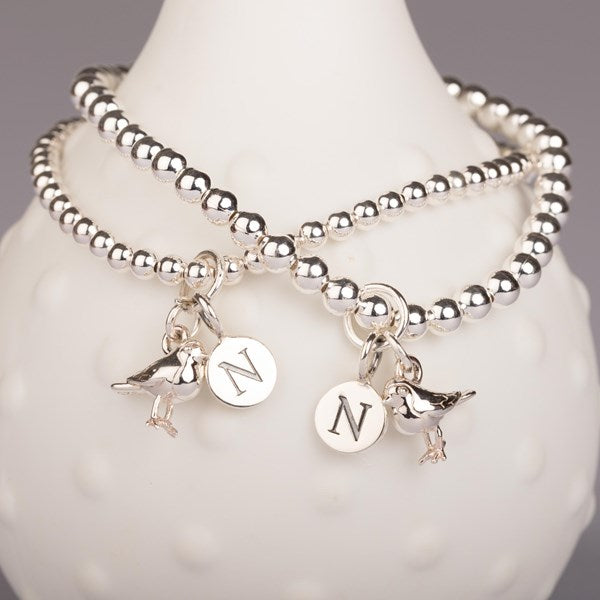 Two silver beaded bracelets displayed on a white textured vase. Each bracelet features a bird charm and a round silver disc charm engraved with the letter 'N'.