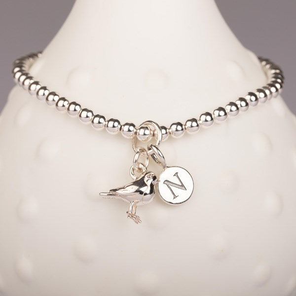 A silver beaded bracelet displayed on a white textured jewelry bust. The bracelet features a small bird charm and a circular initial 'N' charm.