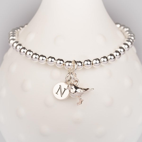 A silver beaded bracelet displayed on a white textured bust, featuring a round engraved disc charm with the letter 'N' and a small bird charm.
