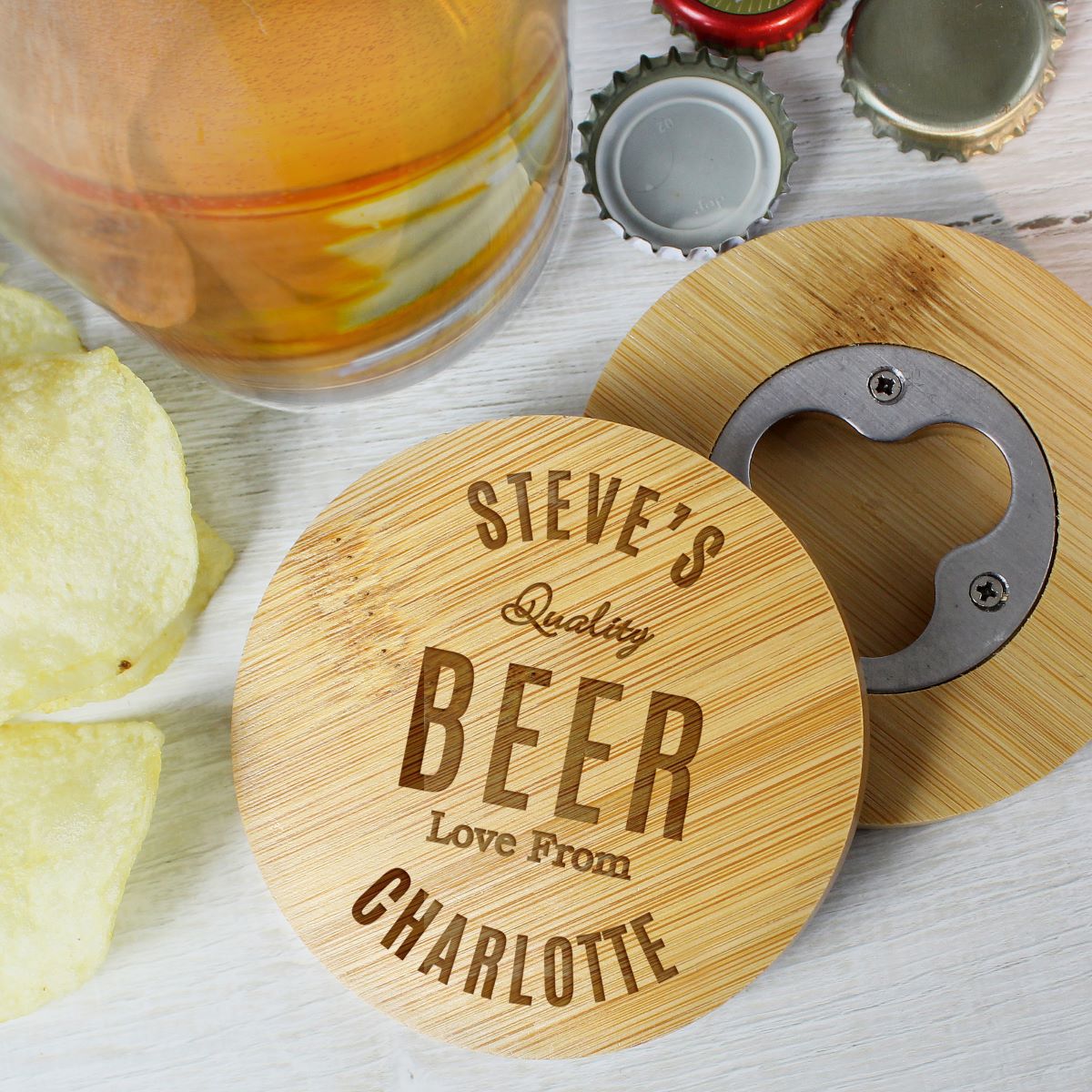 Personalized bamboo beer coasters and bottle openers on a white wooden surface, with a glass of amber beer, potato chips, and bottle caps nearby.
