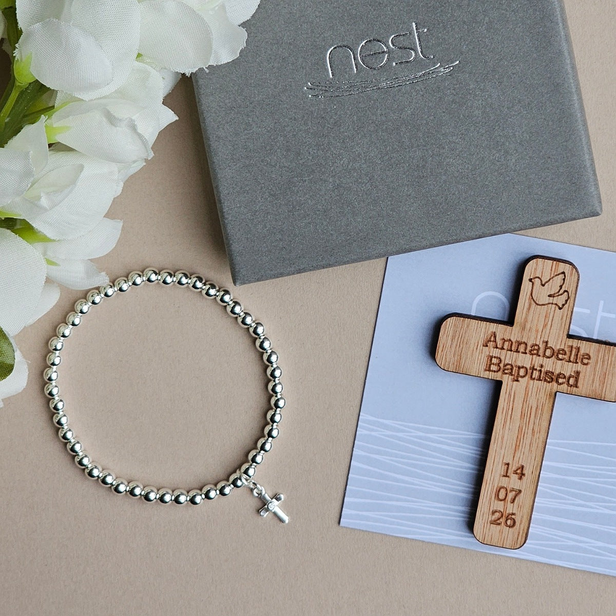 A silver beaded bracelet with a small cross pendant is arranged on a beige surface beside a gray Nest gift box, white flowers, and a wooden baptism cross engraved with the name Annabelle, the date 14/07/26, and a dove motif.