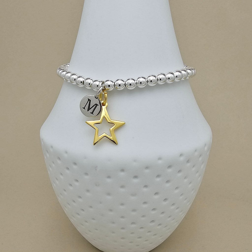 A silver beaded bracelet with a dangling gold star charm and a silver round charm engraved with the letter 'M', displayed on a white textured jewelry bust against a beige background.
