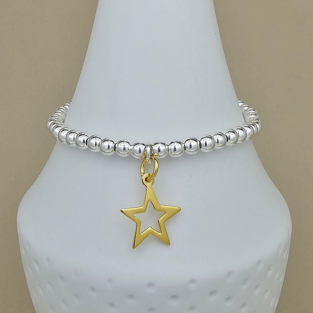 A silver beaded bracelet with a gold star-shaped charm, displayed on a white cone jewelry stand against a beige background.