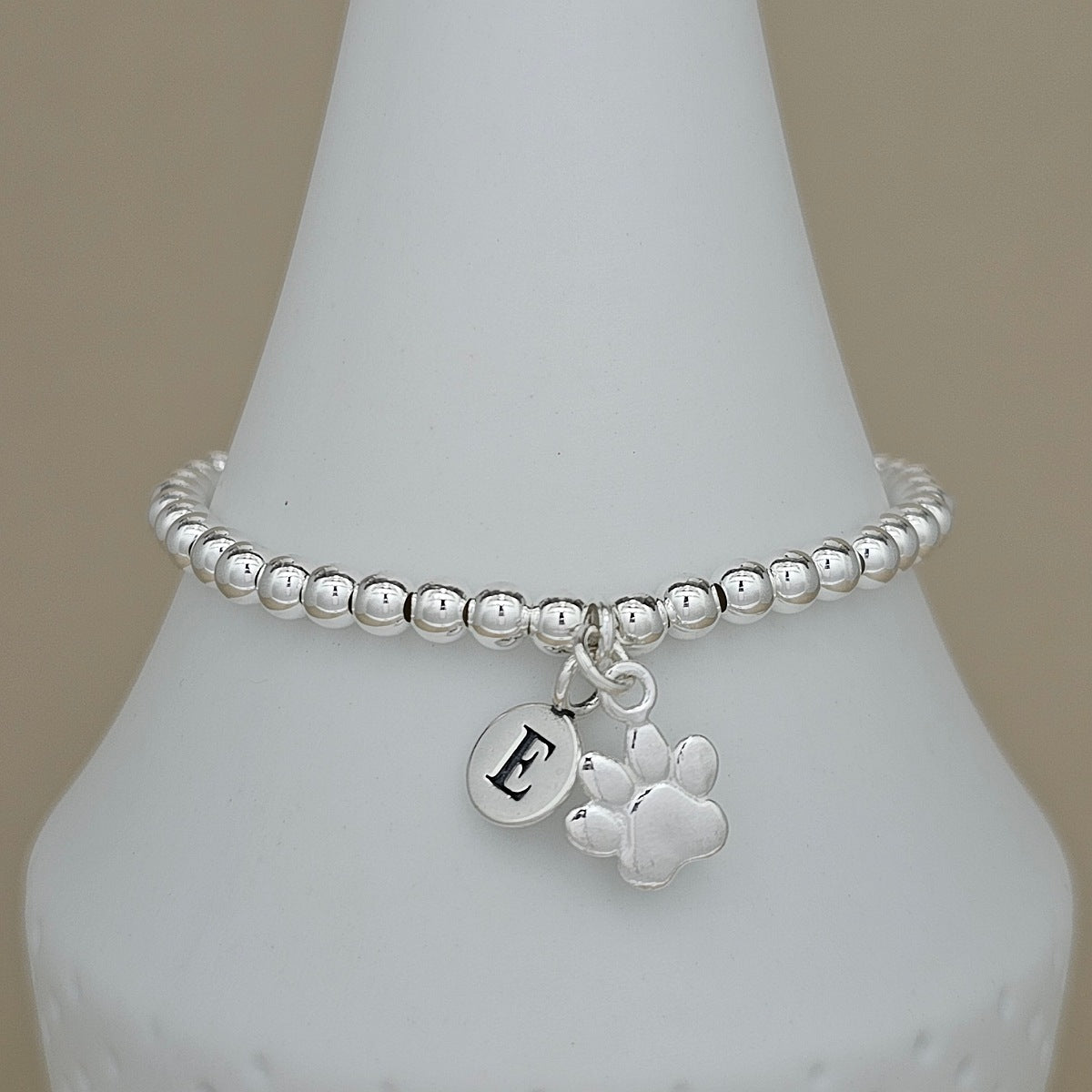 A silver beaded stretch bracelet with a round initial 'E' charm and a paw print charm, displayed on a white cone stand against a neutral beige background.