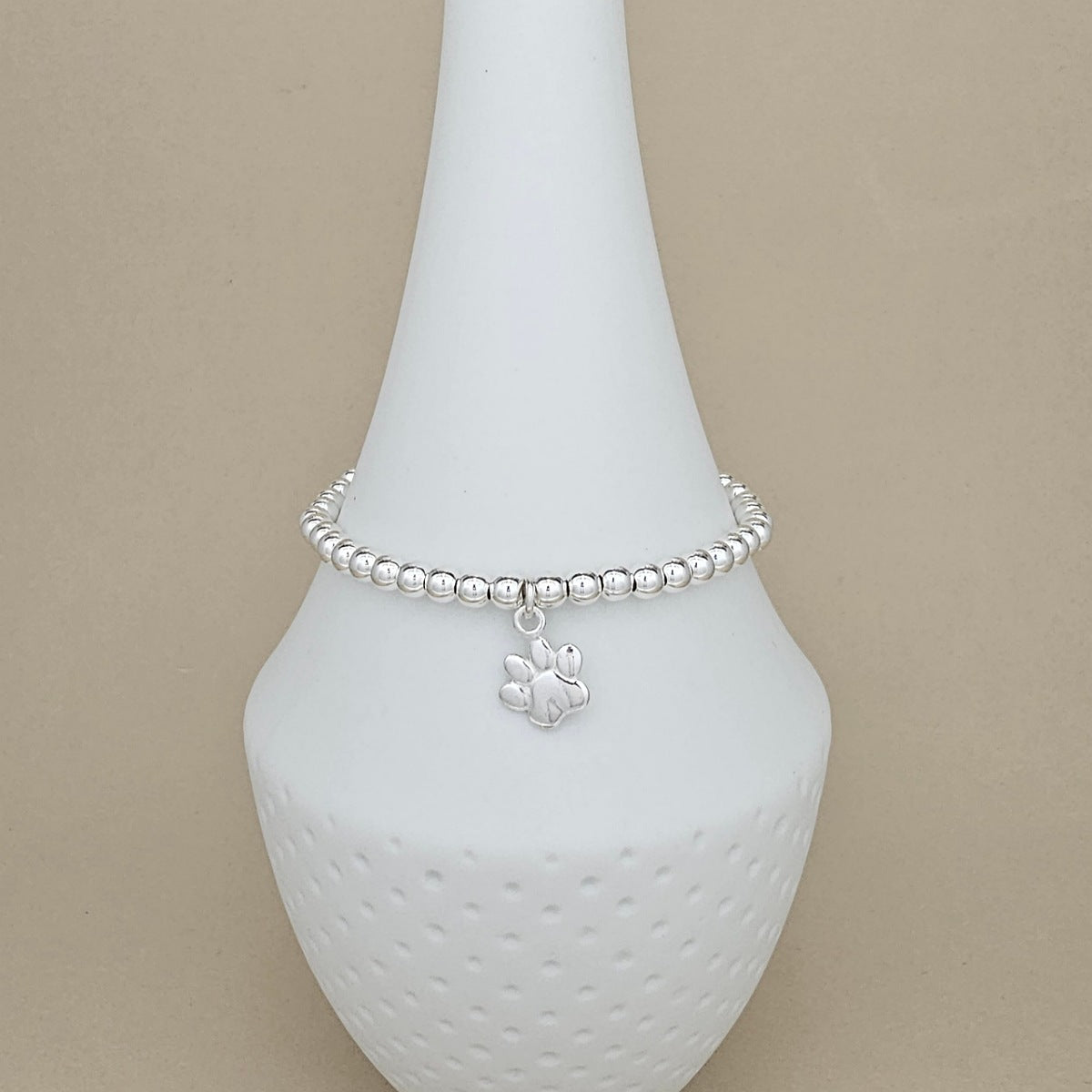 A silver beaded bracelet with a small paw print charm, displayed on a white ceramic jewelry stand against a beige background.