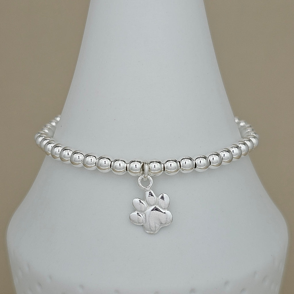 A silver beaded bracelet with a paw print charm, displayed on a white cone-shaped jewelry stand against a neutral beige background.