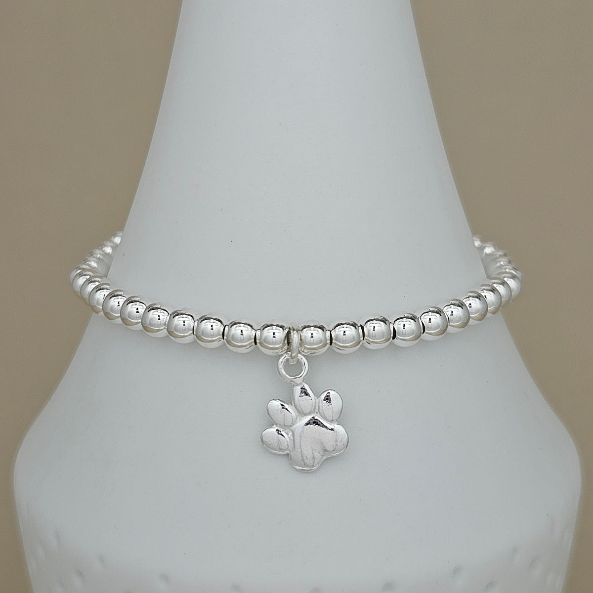 A silver beaded bracelet with a paw print charm, displayed on a white cone-shaped jewelry stand against a neutral beige background.