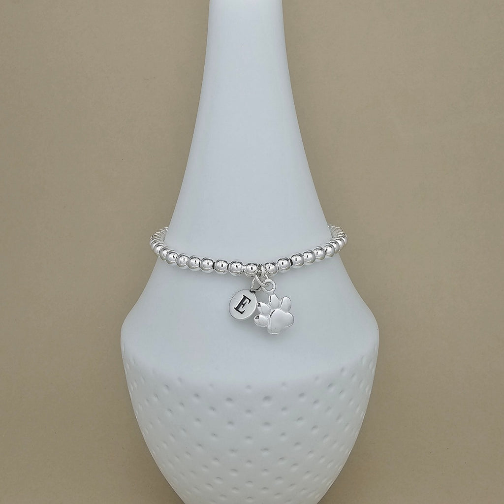 A silver beaded bracelet with a paw print charm and a small initial 'E' charm, displayed on a white ceramic jewelry stand against a beige background.