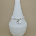 A silver beaded bracelet with a paw print charm and a small initial 'E' charm, displayed on a white ceramic jewelry stand against a beige background.
