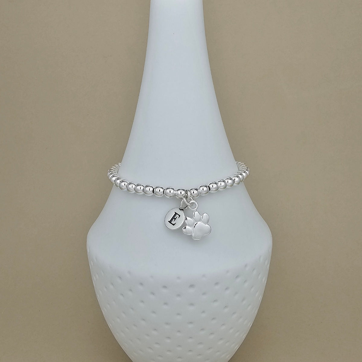 A silver beaded bracelet with a paw print charm and a small initial 'E' charm, displayed on a white ceramic jewelry stand against a beige background.