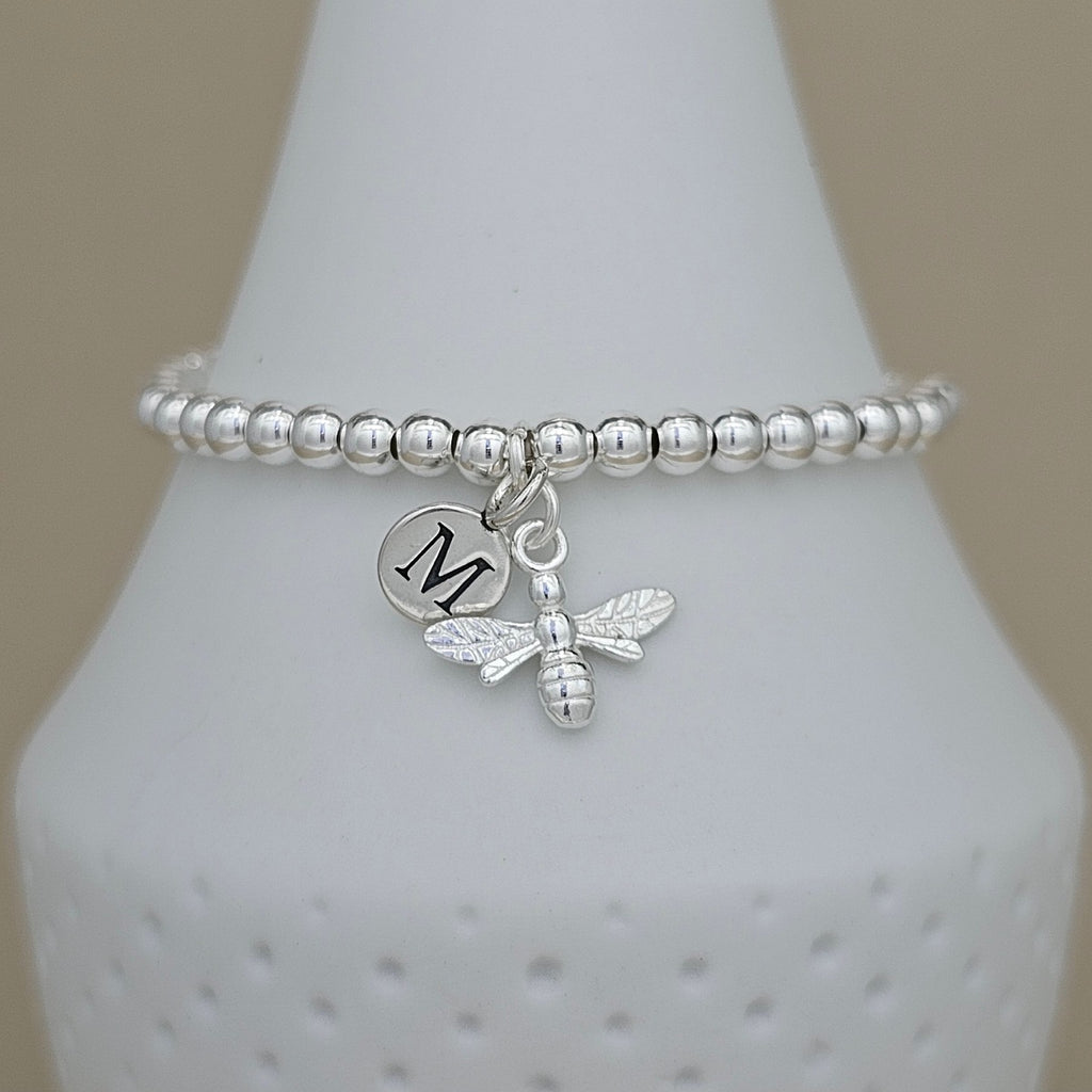 A silver beaded bracelet with a dangling bee charm and a round initial 'M' charm, displayed on a white cone stand against a beige background.