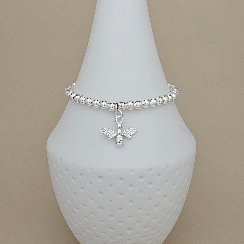 A silver beaded bracelet with a small bee charm, displayed on a white ceramic jewelry stand against a beige background.