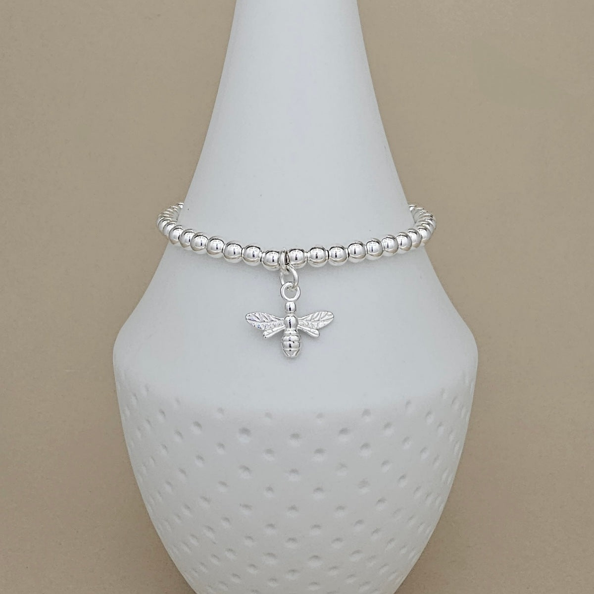 A silver beaded bracelet with a small bee charm, displayed on a white ceramic jewelry stand against a beige background.